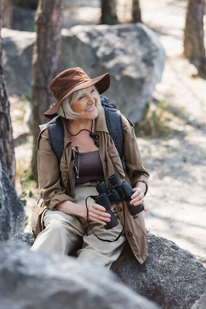 Pozitivní turistka s batohem a dalekohledem sedící na kameni v lese  - Fotografie, Obrázek