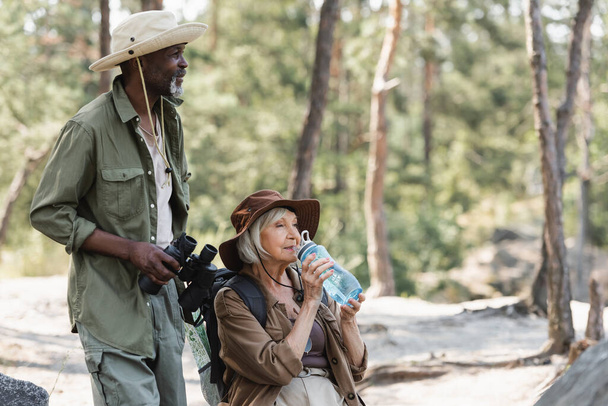 African american tourist holding binoculars near senior wife with water in forest  - 写真・画像