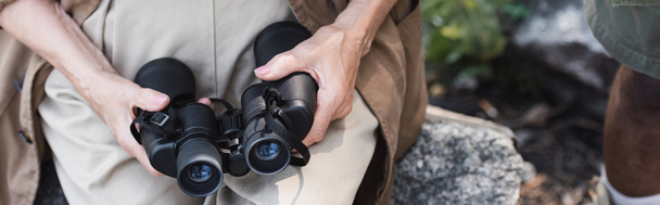 Cropped view of elderly hiker holding binoculars on stone in forest, banner  - Photo, Image