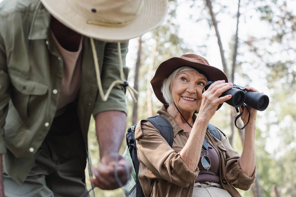 Senior traveler holding binoculars near blurred african american husband in forest  - Photo, Image