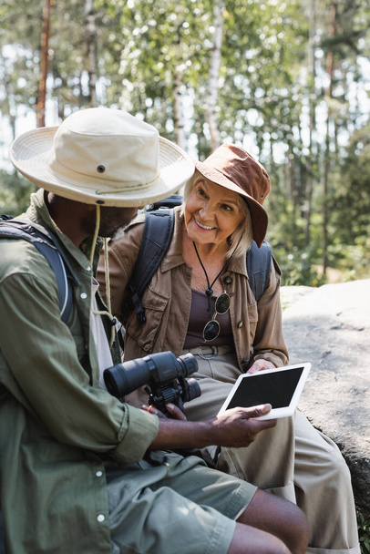 Positive traveler looking at african american husband with binoculars and digital tablet in forest  - Valokuva, kuva