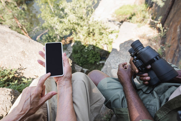 Вид сверху пожилой туристки, указывающей на смартфон рядом с мужем с биноклем на камнях  - Фото, изображение