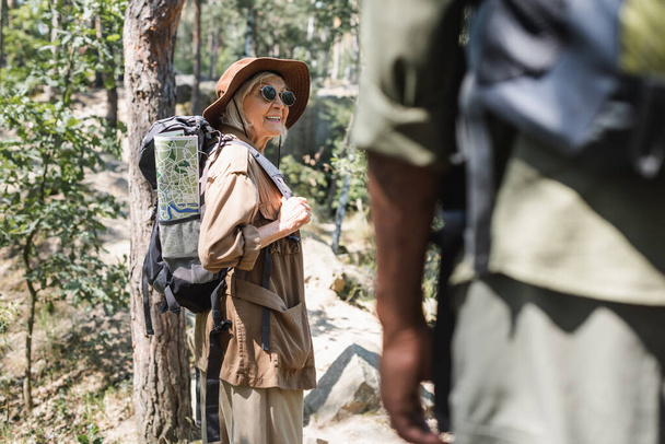 Positive woman with backpack and map standing in forest near blurred african american husband  - Foto, afbeelding