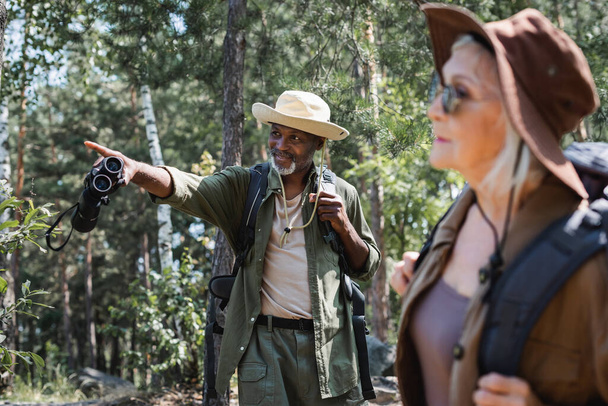 Smiling african american man with binoculars pointing with finger near blurred wife in forest  - Foto, imagen