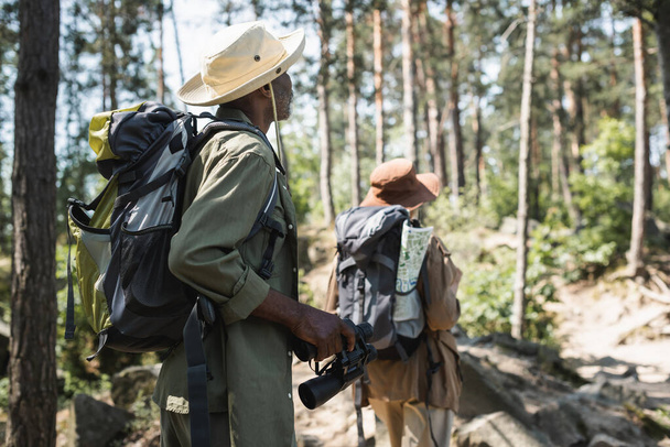Afrykański amerykański turysta z plecakiem i lornetką spacerujący w pobliżu zamazanej żony w lesie  - Zdjęcie, obraz