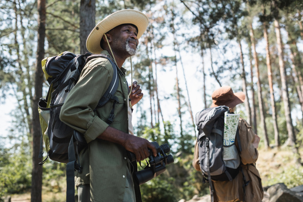 Smiling african american man holding binoculars while hiking with wife in forest  - Zdjęcie, obraz