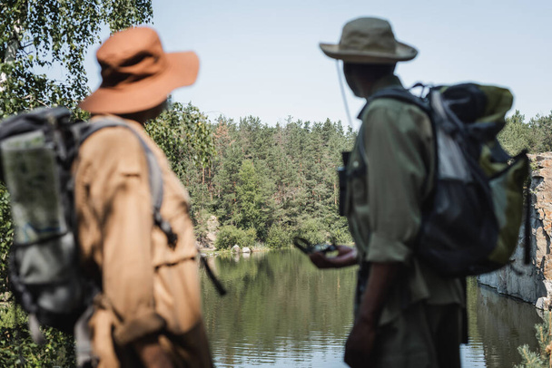 森の近くの湖と前景にバックパックを持つぼやけた異人種間の旅行者  - 写真・画像