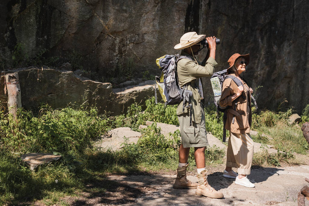 African american man looking though binoculars near wife with backpack and cliff - Photo, Image