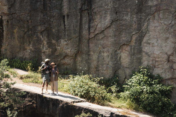Межрасовая старшая пара с рюкзаками, идущая по тропинке рядом со скалой  - Фото, изображение