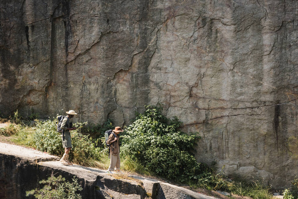 崖の近くのパス上を歩くバックパック付きのシニア多民族カップルのサイドビュー  - 写真・画像