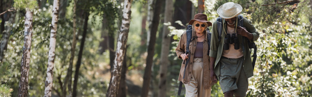 Smiling elderly woman walking near african american husband with binoculars and backpack in forest, banner  - Φωτογραφία, εικόνα