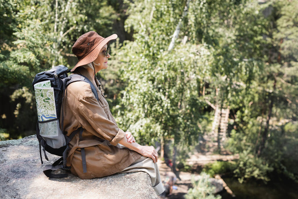 Side view of elderly woman with backpack and map sitting on cliff  - Foto, imagen