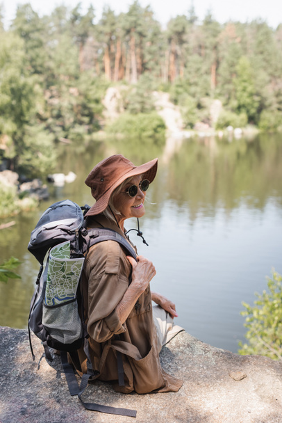 Cheerful senior hiker with backpack and map sitting on cliff near lake  - Foto, Imagem