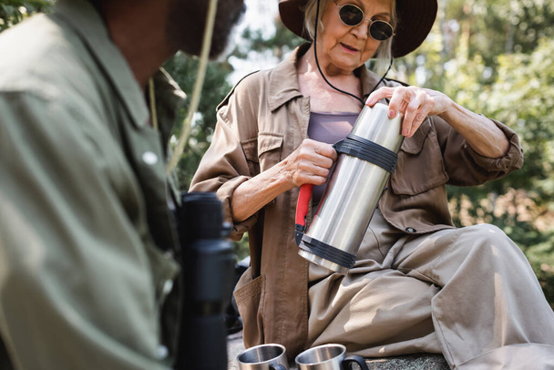 Senior toerist houdt thermoskan in de buurt van kopjes en wazig Afrikaans Amerikaanse man  - Foto, afbeelding