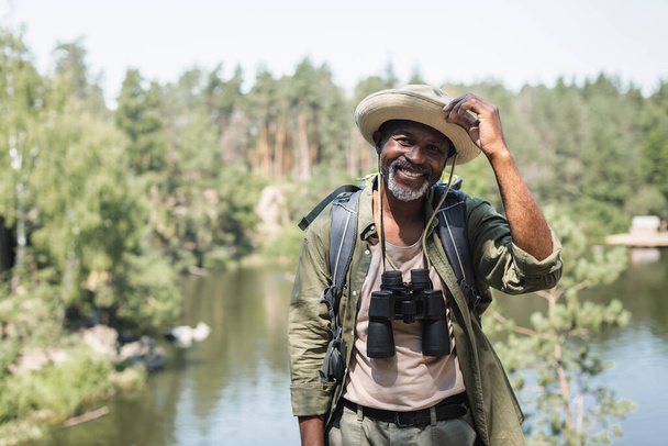 Positive african american hiker with hat and binoculars looking at camera outdoors  - Foto, Imagen