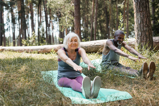 Sportive âgée positive faisant de l'exercice sur un tapis de fitness près d'un mari afro-américain en forêt  - Photo, image