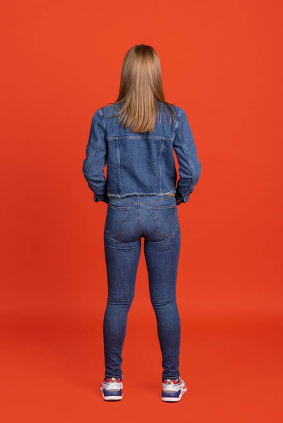 Back view of young red-headed woman standing isolated over red studio background. Copyspace for ad, design. Concept of human emotions, fashion, beauty, youth - Fotografie, Obrázek