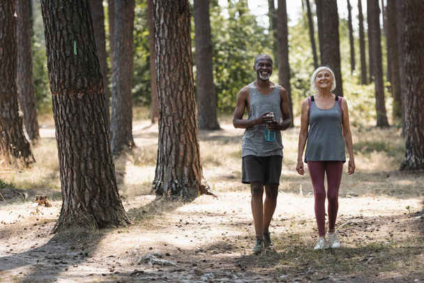 Smiling senior sportswoman walking near african american husband with sports bottle in forest  - 写真・画像