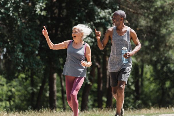 Счастливая многонациональная пара со спортивной бутылкой, указывающей пальцами во время пробежки в парке  - Фото, изображение