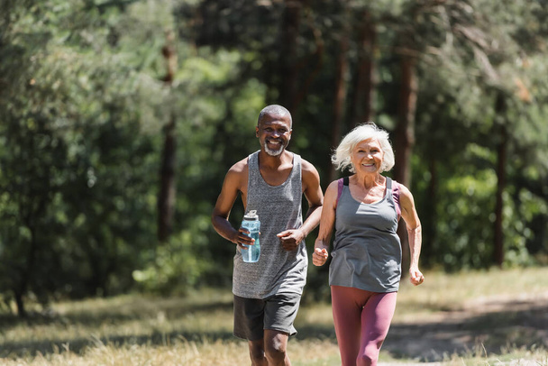 Mosolygó idős sportoló nő fut közelében afro-amerikai férj sport palack az erdőben  - Fotó, kép
