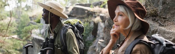 Turista afroamericano senior con bastones de trekking de pie cerca de esposa borrosa en el bosque, pancarta  - Foto, imagen