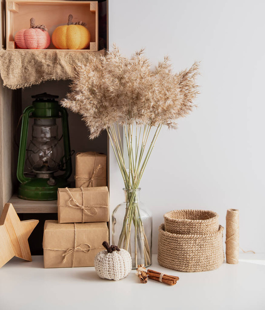 Neutral beige decor colors. Glass vase bottle with a bouquet of dry ears, crocheted jute baskets, pumpkin, gifts in craft paper, wooden star. Simple home decor. - Zdjęcie, obraz