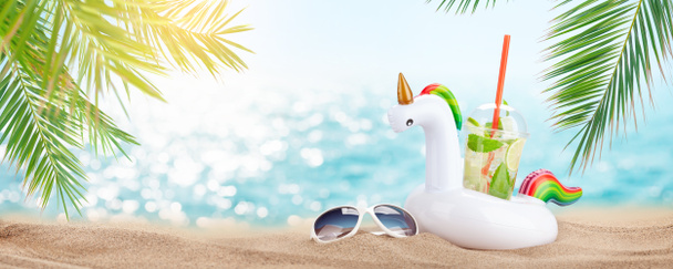 Verse koude mojito cocktail in opblaasbaar eenhoorn speelgoed en zonnebril op tropisch strand met palmen en helder zand. Zomer zee vakantie en reizen concept - Foto, afbeelding