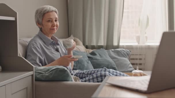 Femme asiatique aux cheveux gris de longueur moyenne portant un pyjama, assise sur le canapé à la maison, buvant dans une tasse, regardant du contenu sur un ordinateur portable - Séquence, vidéo