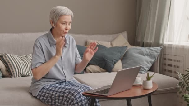 Medium long of older Asian woman wearing pajamas and wireless earphones, sitting on couch in living room, video calling using portable computer - Footage, Video