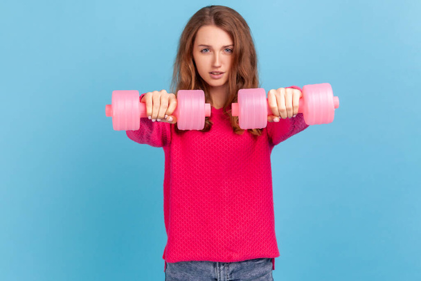 Winsome confident woman with wavy hair wearing pink pullover standing holding out two dumbbells, looking at camera, training her biceps. Indoor studio shot isolated on blue background. - Zdjęcie, obraz