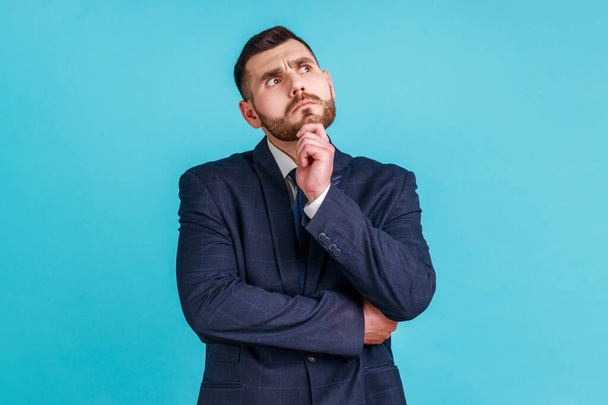Portrait of pensive man wearing official style suit, standing with thoughtful facial expression, holding chin and looking up, being deep in thoughts. Indoor studio shot isolated on blue background. - Foto, afbeelding
