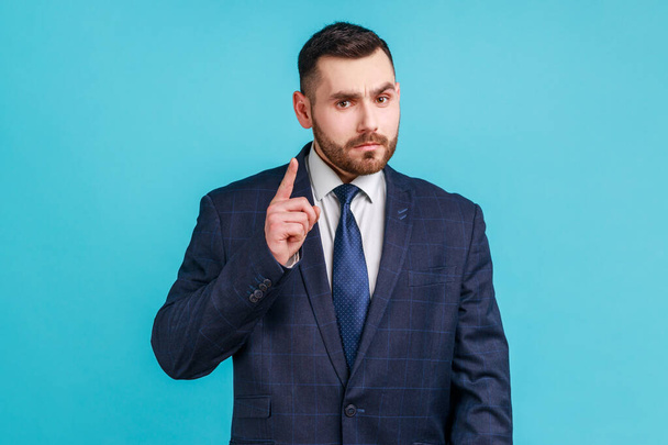 Handsome businessman wearing dark official style suit warning with finger, scolding, strictly looking at camera, pointing finger and alert you. Indoor studio shot isolated on blue background. - Fotoğraf, Görsel