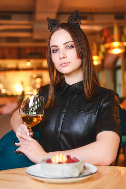 Portrait of young gorgeous woman drinking champagne in glass and looking with smile eating ice cream fruit dessert. Enjoying her leisure time inside a restaurant. Girl in a black dress with cat ears. - Fotoğraf, Görsel