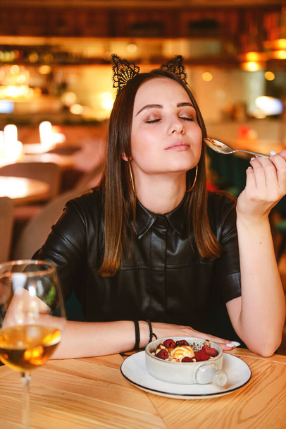 Portrait of young gorgeous woman drinking champagne in glass and looking with smile eating ice cream fruit dessert. Enjoying her leisure time inside a restaurant. Girl in a black dress with cat ears. - Foto, afbeelding