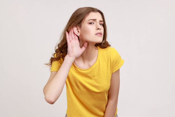 What? I can't hear! Portrait of girl in yellow T-shirt holding hand near ear and listening carefully, having hearing problems, deafness in communication. Indoor studio shot isolated on gray background - Zdjęcie, obraz