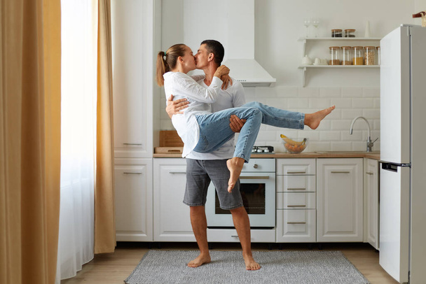 Full length portrait of young man holding his beautiful girlfriend in arms while standing in kitchen at home and kissing her. Both are looking at each other, with love and romantic feelings. - Фото, изображение