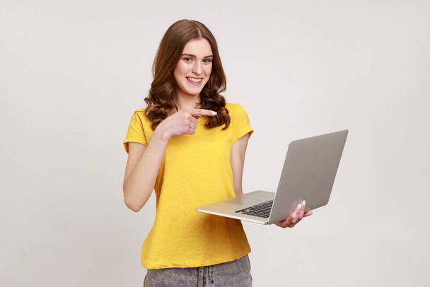 Cheerful positive teenager girl in yellow casual style T-shirt pointing finger at display of laptop, blogger with portable computer. Indoor studio shot isolated on gray background. - 写真・画像