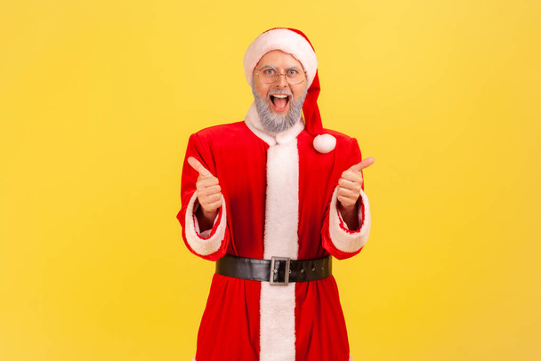 Excited elderly man with gray beard wearing santa claus costume standing with open mouth, screaming happily, showing thumb up to camera. Indoor studio shot isolated on yellow background. - Valokuva, kuva