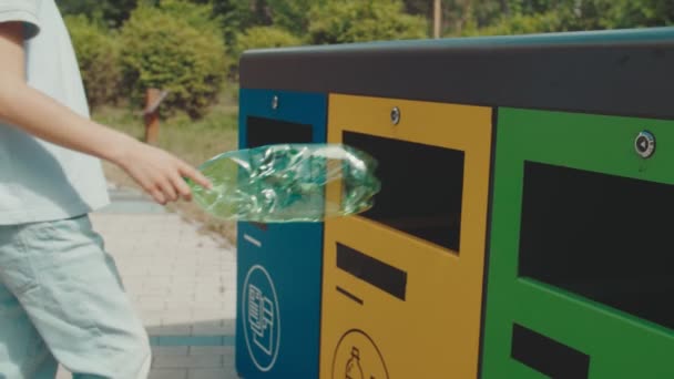 Kędzierzawy chłopiec wyrzucając pustą plastikową butelkę do kosza na śmieci na zewnątrz - Materiał filmowy, wideo