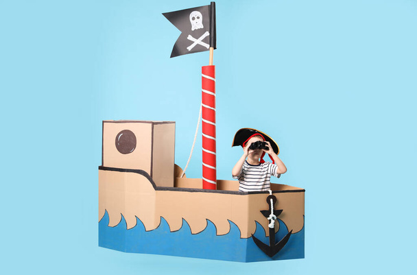 Little boy playing with binoculars in pirate cardboard ship on turquoise background - Fotó, kép