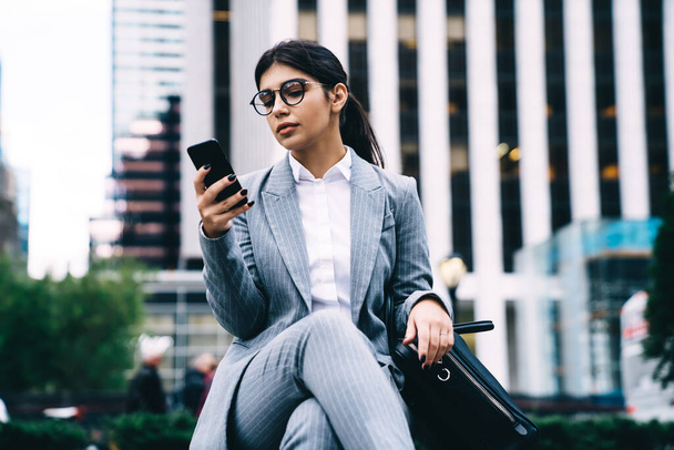 Latino businesswoman reading financial news on web page via cell smartphone, confident female CEO using mobile phone for search contact number or data info during work break at urban setting - Фото, изображение