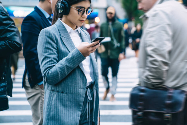 Crop thoughtful woman in glasses wearing office clothes attentively using smartphone while standing on busy city street in wireless headphones - 写真・画像