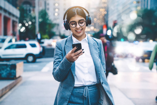 Brunette businesswoman in formal clothing with round glasses using smartphone and listening to music while walking in downtown on blurred background - Foto, afbeelding