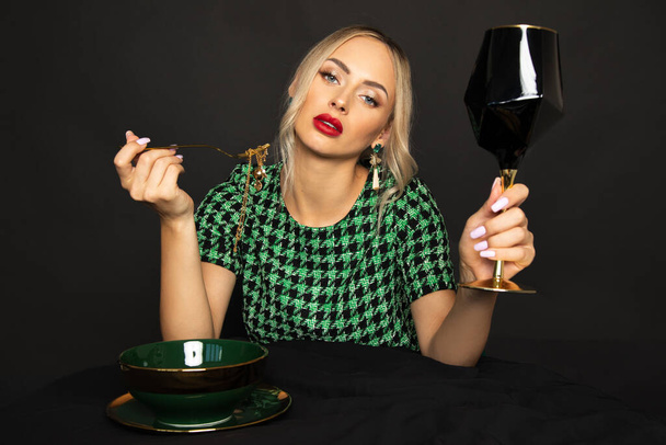 gorgeous blonde woman looks down at camera on black background with glass of wine at table set with expensive dishes. Luxury lifestyle - Photo, Image