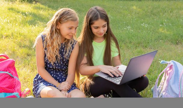 Twee meisjes met laptop en aktetassen die huiswerk maken of plezier hebben terwijl ze buiten op het gras zitten - Foto, afbeelding