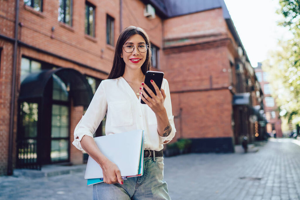 Half length portrait of cheerful Spanish woman designer with creative sketchbook in hand posing at city street during free time, positive hipster girl in optical eyewear using 4g connection - Фото, изображение