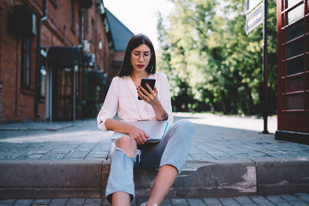 Concentrated slim young lady in glasses in white shirt ripped jeans sitting on curb near telephone booth in street texting on mobile phone holding folders in hands - Foto, Imagem