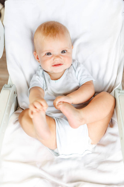 Cute little noworodka dziewczyna z uśmiechniętą twarzą patrząc w aparat na białym tle. Niemowlę odpoczywające bawiące się na łóżeczku w domu. Matczyna koncepcja szczęśliwego dziecka. - Zdjęcie, obraz