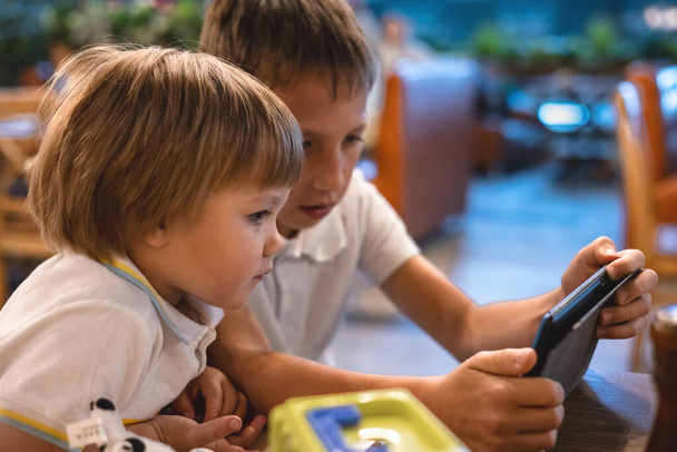 Boy and toddler playing video games on tablet online on internet while waiting orders in restaurant. Children using smartphone. Kids using technology. Screen time addiction. - Foto, Imagen