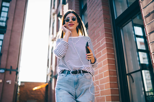 From below of fashionable brunette in trendy casual outfit and sunglasses smiling while using earphones with mobile phone and looking at camera at city street  - Photo, Image